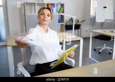 Femme d'affaires stricte assise à la réception et donnant le document à quelqu'un Banque D'Images
