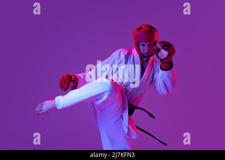 Photo en studio d'un chasseur de taekwondo mâle en uniforme sportif en action isolé sur fond violet dans la lumière de néon. Concept de sport, d'entraînement Banque D'Images