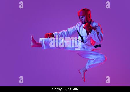 Photo en studio d'un chasseur de taekwondo mâle en uniforme sportif en action isolé sur fond violet dans la lumière de néon. Concept de sport, d'entraînement Banque D'Images