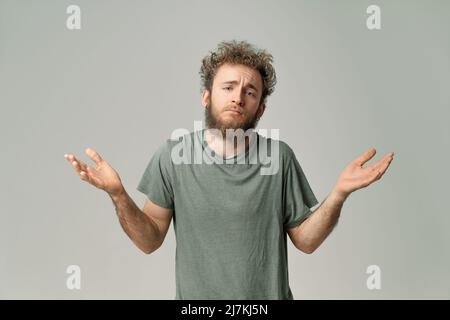 Gesturant Je sais que ou je suis DÉSOLÉ jeune beau barbu sauvage cheveux bouclés homme avec des yeux bleu vif isolé sur fond gris. Jeune homme pensant en t-shirt vert sur blanc. Copier l'espace. Banque D'Images