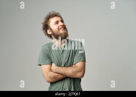 Jeune homme souriant, avec des cheveux bouclés et sauvages, des yeux bleu vif qui regardent avec les mains repliées isolés sur fond blanc. Jeune homme pensant dans un t-shirt vert sur fond blanc. Banque D'Images