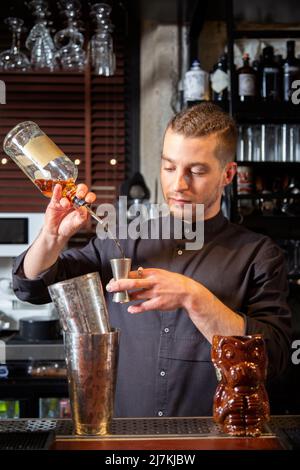 Jeune barman à la coiffure tendance en uniforme en versant ron dans un puzzle tout en se tenant au comptoir et en préparant un cocktail d'alcool au bar Banque D'Images