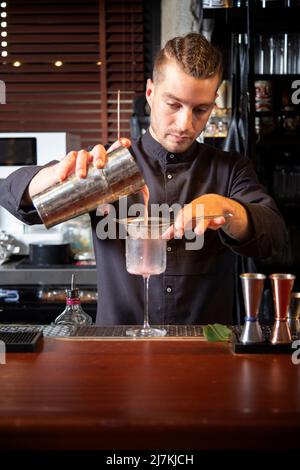 Jeune barman concentré en uniforme noir, en versant un cocktail exotique dans une passoire tout en se tenant au comptoir dans un bar moderne Banque D'Images