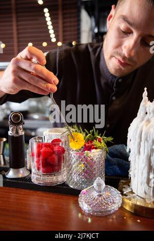 Un barkeeper sérieux en uniforme noir décorant un cocktail avec une fleur fraîche pendant le travail au bar Banque D'Images