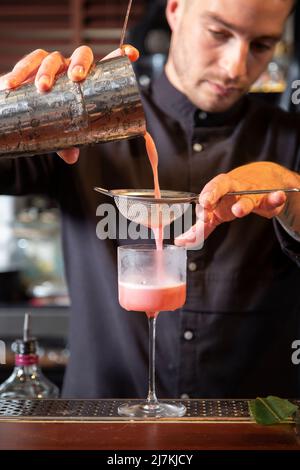 Jeune barman concentré en uniforme noir, en versant un cocktail exotique dans une passoire tout en se tenant au comptoir dans un bar moderne Banque D'Images