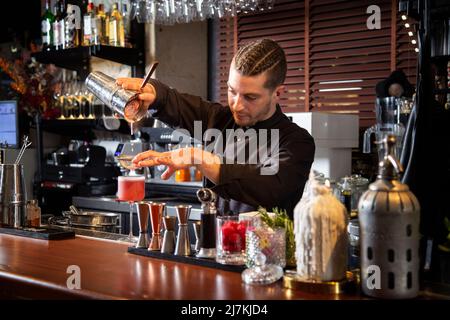 Jeune barman concentré en uniforme noir, en versant un cocktail exotique dans une passoire tout en se tenant au comptoir dans un bar moderne Banque D'Images