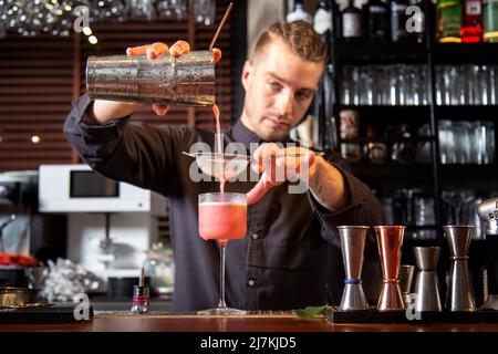 Jeune barman concentré en uniforme noir, en versant un cocktail exotique dans une passoire tout en se tenant au comptoir dans un bar moderne Banque D'Images