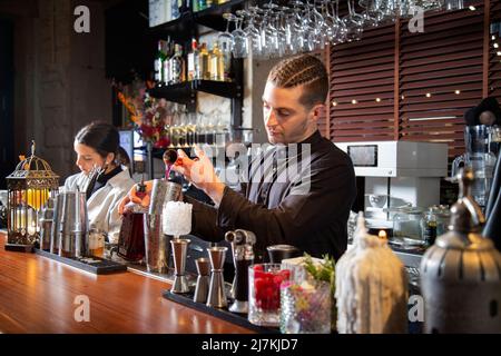 Jeunes hommes et femmes barmen concentrés en uniforme préparant divers cocktails alcoolisés tout en se tenant au comptoir dans un bar moderne Banque D'Images