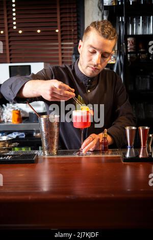 Un jeune barkeeper élégant avec cheveux tressés en uniforme noir décorant un délicieux cocktail rose avec des feuilles fraîches pendant le travail au bar Banque D'Images