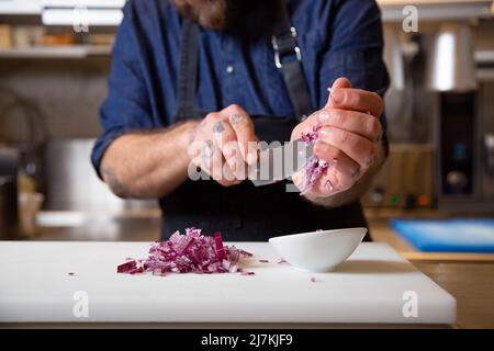 Crochez un chef masculin tatoué et méconnu dans un tablier en mettant un oignon rouge haché avec un couteau dans un bol tout en cuisinant dans une cuisine légère Banque D'Images