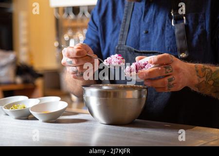 Un petit chef de cuisine tattooed coupé en uniforme en mettant de l'oignon rouge frais dans un bol tout en travaillant à table dans la cuisine légère du restaurant Banque D'Images