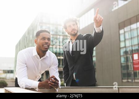 Content collègues masculins multiraciaux en tenue habillée pointant et regardant loin tout en se tenant près du bâtiment de bureau moderne le jour ensoleillé Banque D'Images