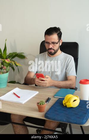 Homme concentré dans les écouteurs messagerie texte sur smartphone et de faire de la paperasserie pendant le travail à table dans le bureau à domicile Banque D'Images