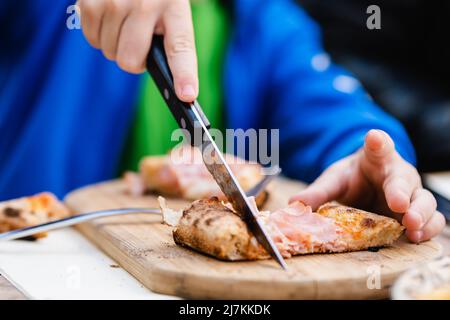 Attention douce de l'enfant méconnaissable dans des vêtements décontractés en coupant de délicieux jambon pizza tout en prenant le déjeuner dans le restaurant de rue Banque D'Images