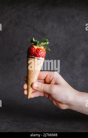 Main de la récolte personne méconnaissable tenant un savoureux cône de glace douce avec fraise rouge mûre sur fond gris en studio Banque D'Images