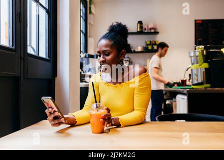 Jolie femme afro-américaine souriante dans un chemisier jaune vif une tasse de jus de fruit jetable et la navigation sur internet sur un smartphone assis Banque D'Images
