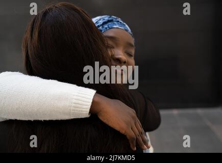 Jeune femme afro-américaine positive en tenue décontractée et foulard souriant et embrassant une amie anonyme dans la rue Banque D'Images