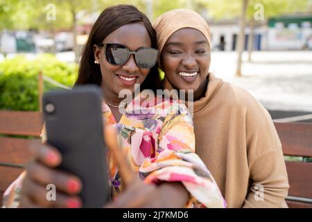 Jeune femme afro-américaine joyeuse dans des lunettes de soleil tendance souriant et prenant selfie sur smartphone avec une amie musulmane heureuse assise sur le banc Banque D'Images