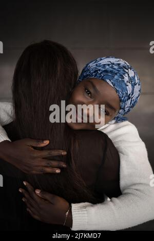 Jeune femme afro-américaine positive en tenue décontractée et foulard souriant et embrassant une amie anonyme dans la rue Banque D'Images