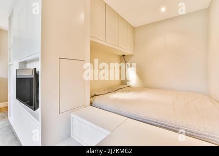 Lit blanc confortable placé au mur près des armoires avec lampe lumineuse placée dans une chambre spacieuse et lumineuse avec télévision moderne dans l'appartement Banque D'Images