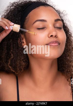 Magnifique jeune modèle hispanique féminine avec une peau parfaite et de longs cheveux bouclés application d'acide hyaluronique sur le visage avec les yeux fermés pendant la procédure de beauté Banque D'Images