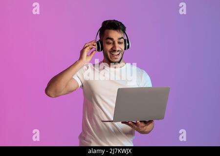 Un homme arabe enthousiaste portant un casque, regardant l'écran d'ordinateur portable, a surjoyé à propos de l'énorme vente dans le magasin Web dans la lumière de néon Banque D'Images