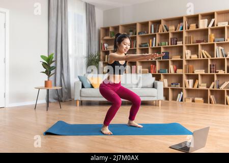 Jeune femme asiatique de forme s'accroupir sur un tapis de fitness devant un ordinateur portable, s'entraîner en ligne à la maison, espace libre Banque D'Images