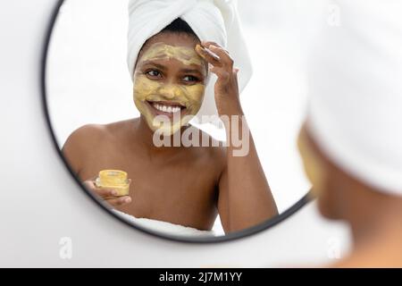 Souriante afro-américaine femme adulte de taille moyenne regardant dans le miroir et appliquant le paquet de visage Banque D'Images