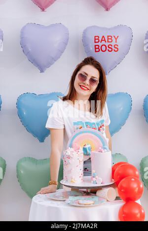 Belle jeune femme en lunettes de soleil avec grand gâteau d'anniversaire sur de nombreux ballons de coeur colorés fond. Sourires, drôle jour de Valentines, fête d'anniversaire. Banque D'Images