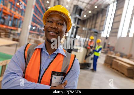 Portrait d'un jeune contremaître souriant et confiant portant un casque avec les bras croisés debout dans l'entrepôt Banque D'Images