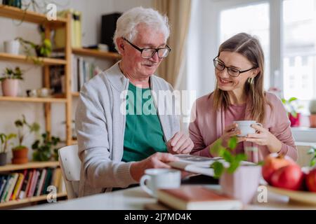 Fille adulte rendant visite à son père senior à la maison et prenant un café ensemble, regardant un livre. Banque D'Images