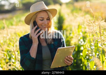 Souriante femme casienne de taille moyenne adulte portant un chapeau tenant un Tablet pc et parlant au-dessus d'un smartphone à la ferme Banque D'Images