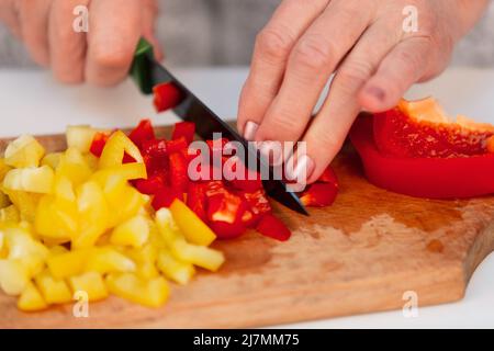 Les mains de la vieille femme ont coupé des poivrons rouges et jaunes sur une planche à découper avec un couteau en céramique Banque D'Images