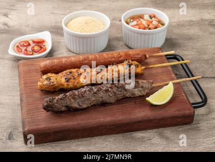 Brochettes de viande, poulet et saucisse de porc sur une planche de bois avec farofa et vinaigrette. Banque D'Images