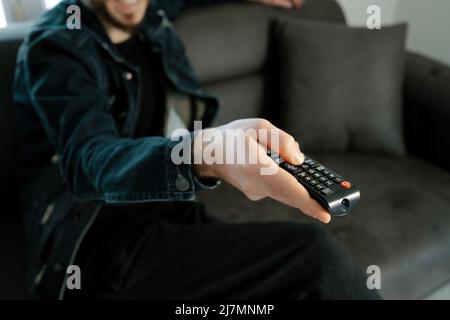 Télécommande de télévision, jeune homme assis sur un canapé dans le salon changer de chaînes avec télécommande en main, services de streaming vidéo et sites de visionnement de séries TV Banque D'Images