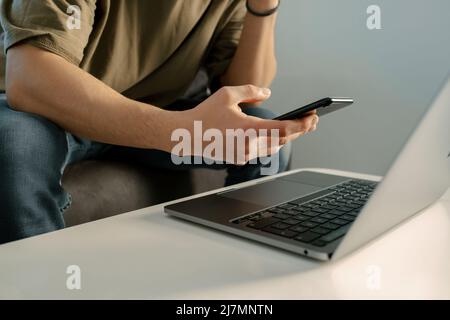 Téléphone à portée de main, homme adulte tient son téléphone, ordinateur portable est sur la table et il parcourt les sites de réseautage social, jeune homme vérifie ses mails Banque D'Images