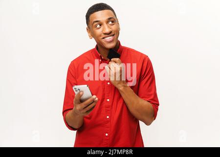 Homme afro-américain souriant en chemise rouge isolé sur fond blanc pensant et rêvant d'acheter longtemps attendu tenant la carte de crédit et le smartphone entre les mains, prêt à payer en ligne Banque D'Images