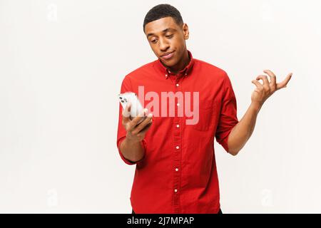 Confus jeune Afro-américain court-coupé dans la chemise rouge tient le smartphone et les épaules isolées sur fond blanc, déçu homme multiracial regarde l'écran du téléphone Banque D'Images