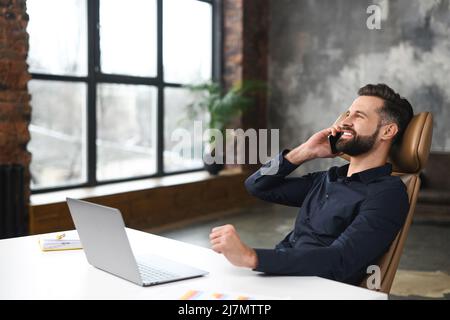 Décontracté caucasien gars assis au bureau et utilisant un ordinateur portable moderne pour un travail. Le jeune homme parle sur un smartphone et sourit, tout en ayant une conversation téléphonique avec des clients ou des collègues Banque D'Images