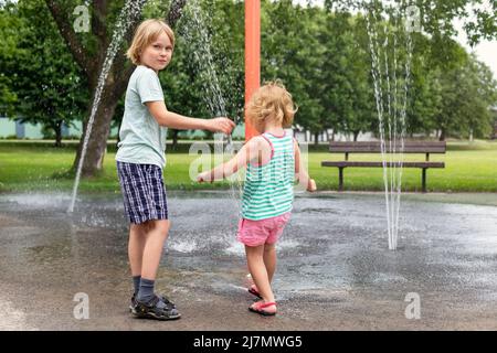 Les petits enfants jouent à la fontaine d'eau dans l'aire de jeux du parc le jour d'été chaud et ensoleillé. Des enfants heureux qui s'amusent à l'extérieur en été. Banque D'Images