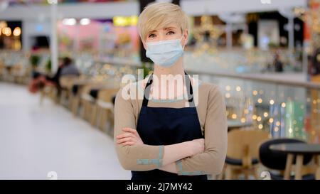 Adulte adulte mature moyenne d'âge 40s femme dans un masque médical restaurant employé de café regardant la caméra croisant les bras femme serveuse vendeuse en tablier travaillant Banque D'Images