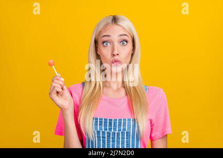 Portrait d'une jolie petite fille adolescente girly léchante bonbon avec les yeux piquant les lèvres isolées sur fond jaune vif Banque D'Images