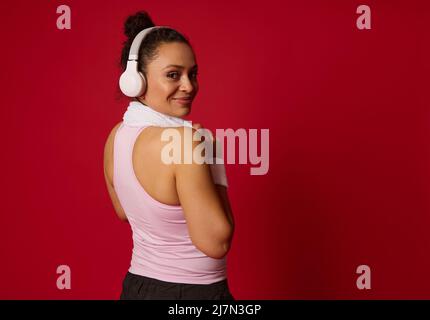 Charmante femme active afro-américaine mature dans les vêtements de sport, casque sans fil, sourires, regardant les épaules à l'appareil photo, se détend après l'entraînement, iso Banque D'Images
