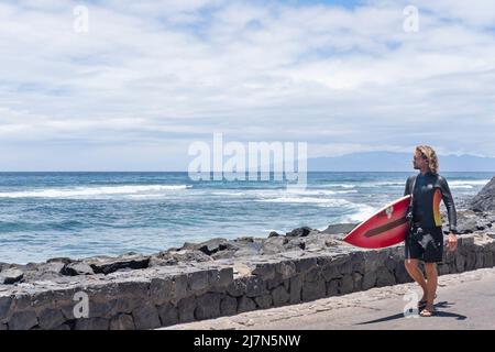 Jeune surfeur mâle portant une combinaison et tenant une grande planche de surf tout en marchant sur la promenade de Las Americas, Tenerife, îles Canaries, Espagne Banque D'Images