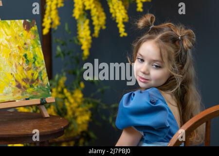 Une fille est assise près d'un mimosa et peint une image dans une robe bleue mimosa brosse bouquet cheveux, pour le portrait assis pour l'enfance pour sourire heureux, joie Banque D'Images