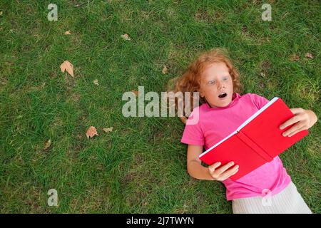 Choqué petite fille redhead lisant le livre sur l'herbe verte Banque D'Images