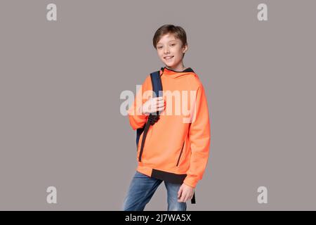 Adolescent de race blanche dans un sweat-shirt orange avec sac à dos sur son dos posé en studio sur fond gris. Joyeux étudiant souriant nouveau jour Banque D'Images