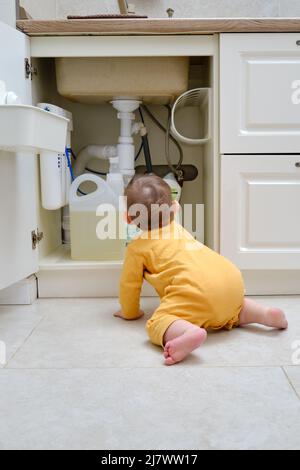 Bébé garçon joue avec des détergents et des produits de nettoyage dans une armoire de cuisine ouverte. Problèmes de sécurité des enfants dans la chambre d'accueil, petit enfant Banque D'Images