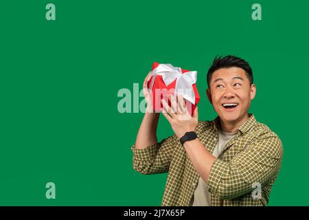 Devinez ce qui se trouve à l'intérieur. Un homme asiatique mûr et heureux écoute ce qui vibre dans une boîte cadeau et regarde de côté l'espace libre Banque D'Images