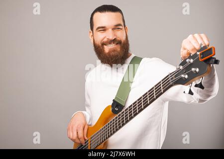 Un homme barbu souriant tient une guitare basse en bois dans un studio sur fond gris. Banque D'Images